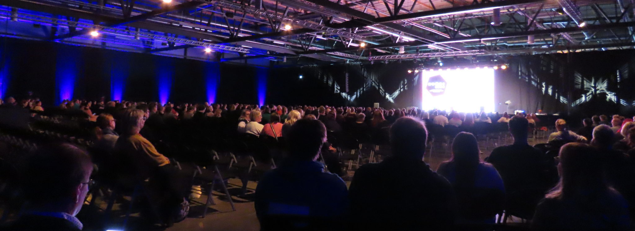 Iso sisätila, jossa taustalla esiintymislava jossa iso näyttökangas, ihmisiä selkäpuolelta lukemattomissa riveissa, katossa valokalustoa ja salin valaistus hämärä.