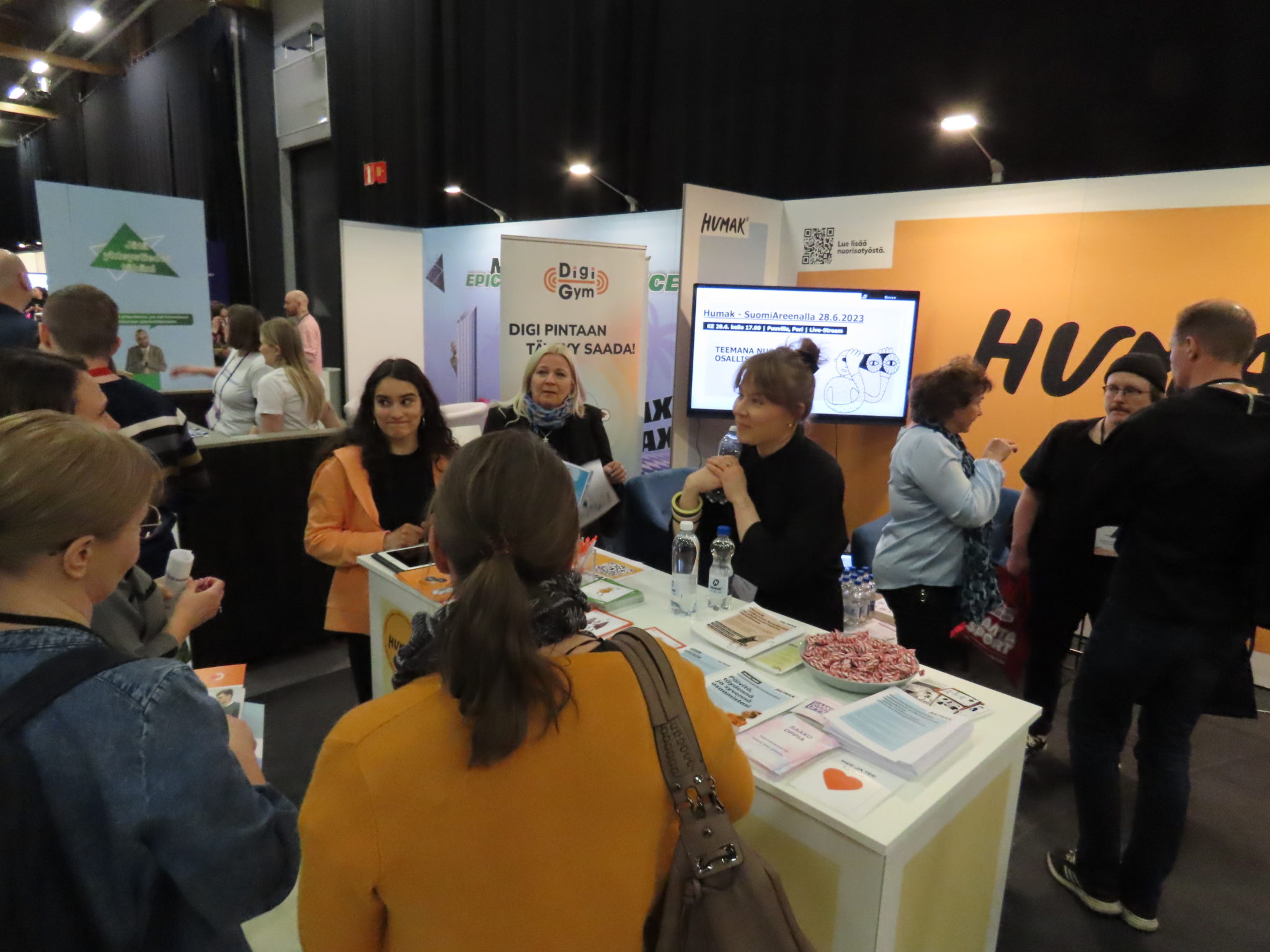 Messuosasto, jossa ihmisiä esittelypöydän ympärillä. Pöydällä erilaisia esitteitä ja makeisia. Takaseinässä lukee Humak ja reunassa roll-up, jossa lukee Digi Gym.