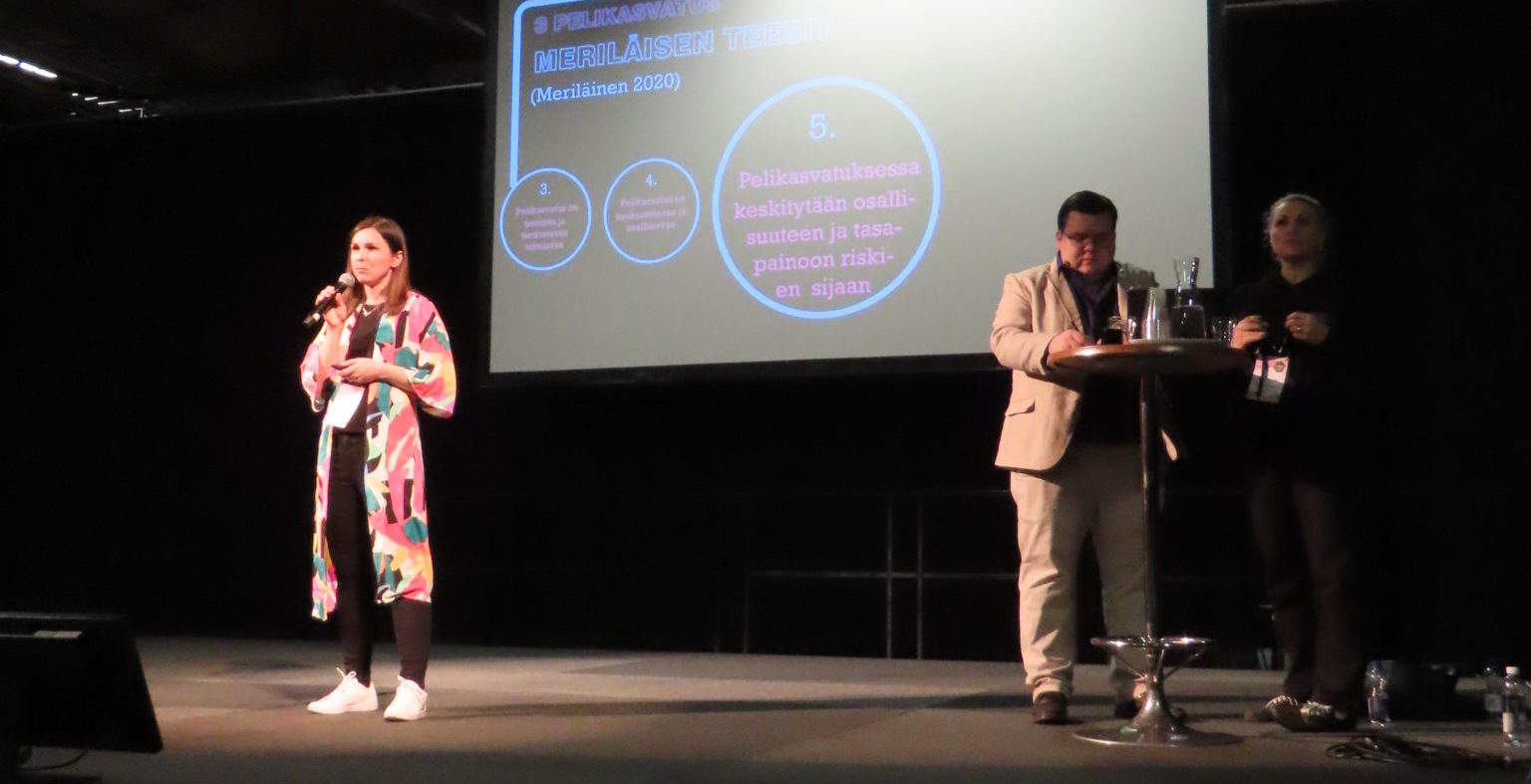 Sisätilassa oleva esiintymislava, jossa kolme ihmistä. Yksi nainen puhuu mikrofoniin, taustalla laajakangas johon heijastettu tekstiä. 