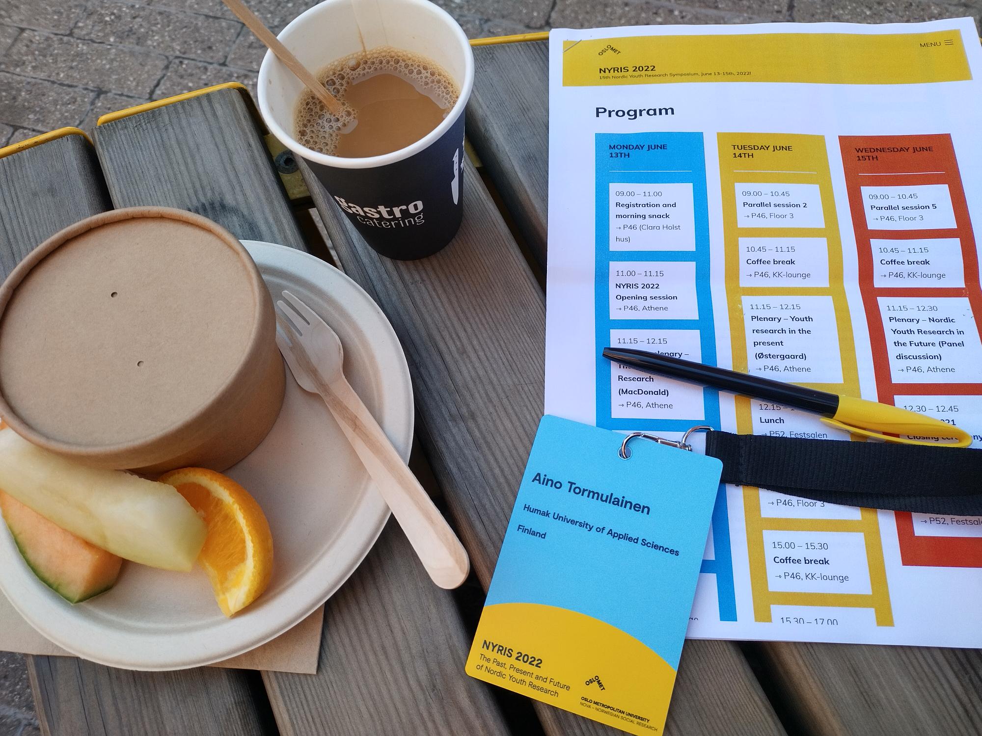 Hedelmiä lautasella, kahvi pahvimukissa, osallistujan nimilappu sekä konferenssin aikataulu paperilla.