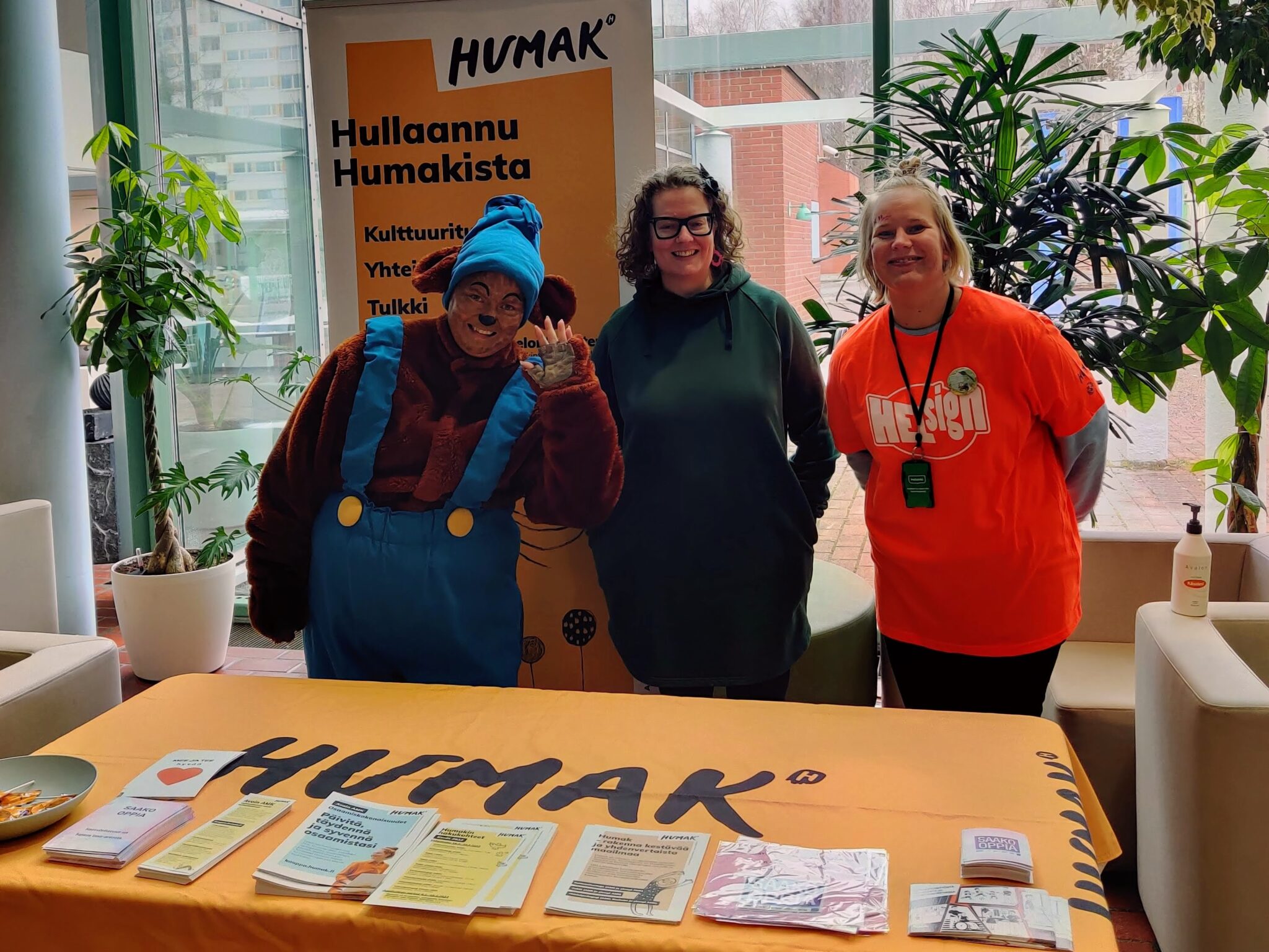 Humakin oranssin esittelypöydän takana seisoo hymyillen lehtori ja kaksi tulkkiopiskelijaa, joista toinen on pukeutunut nalleksi.