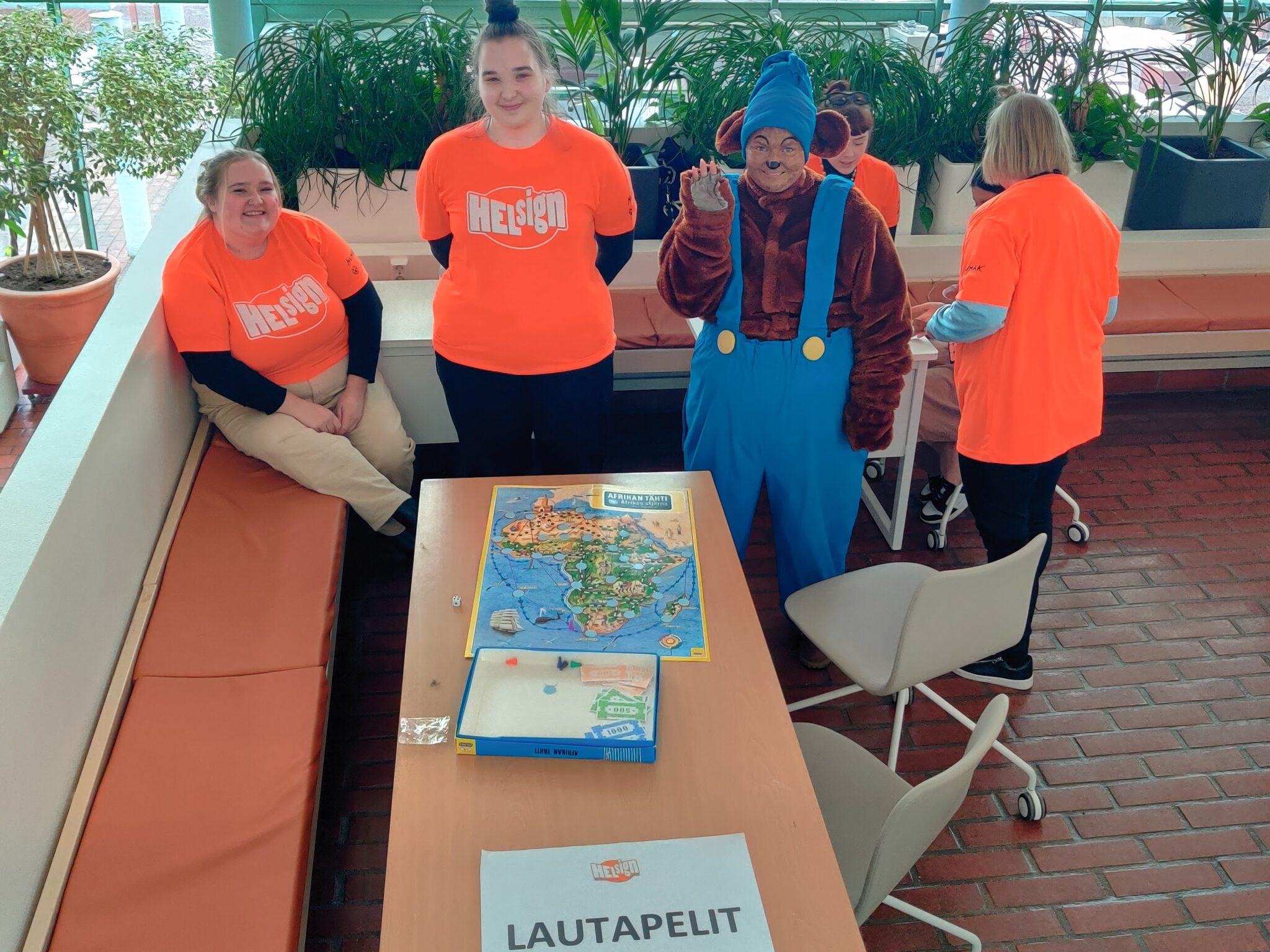  Lautapelipisteen pöydällä on Afrikan tähti -peli. Pöydän takana on kolme tulkkiopiskelijaa, joista yksi on pukeutunut nalleksi. Taustalla kaksi tulkkiopiskelijaa neuvoo nuorta pelaamisessa. 