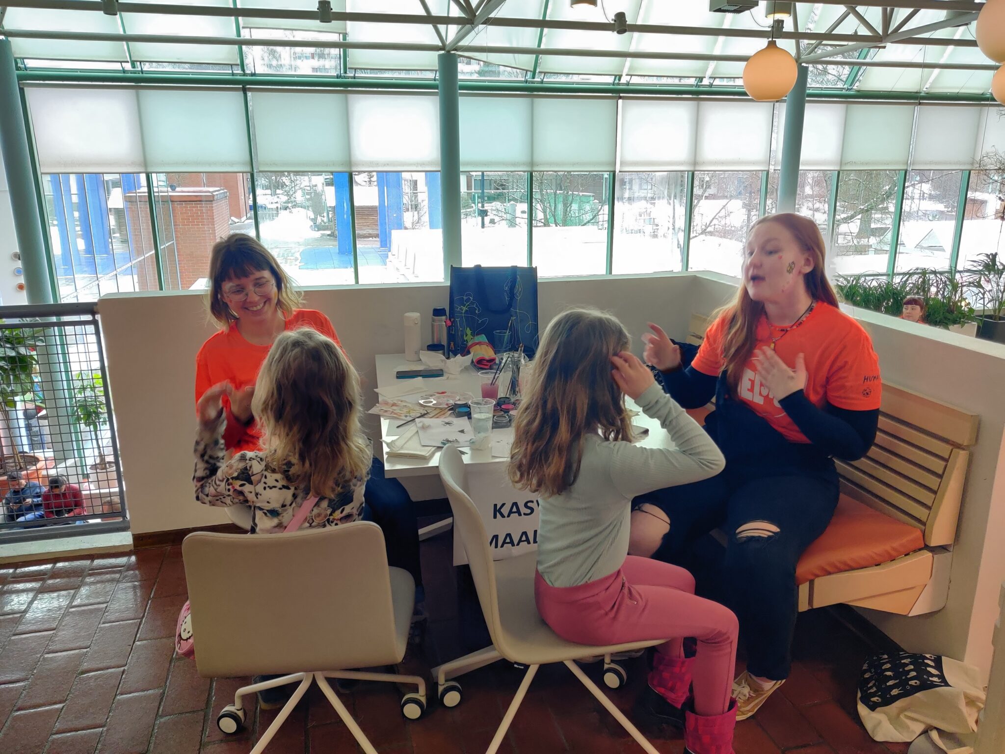 Kasvomaalauspisteessä kaksi oranssipaitaista opiskelijaa maalaa kahden lapsen poskeen kuvaa. Pöydällä on maaleja ja siveltimiä.