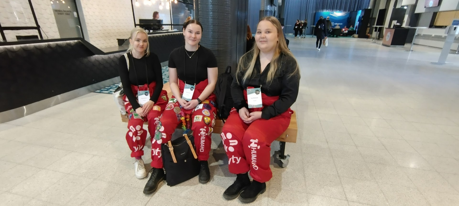 Kolme opiskelijahaalareihin pukeutunutta nuorta naista istuu penkillä tapahtumahallin sisällä ja hymyillee kameralle. 
