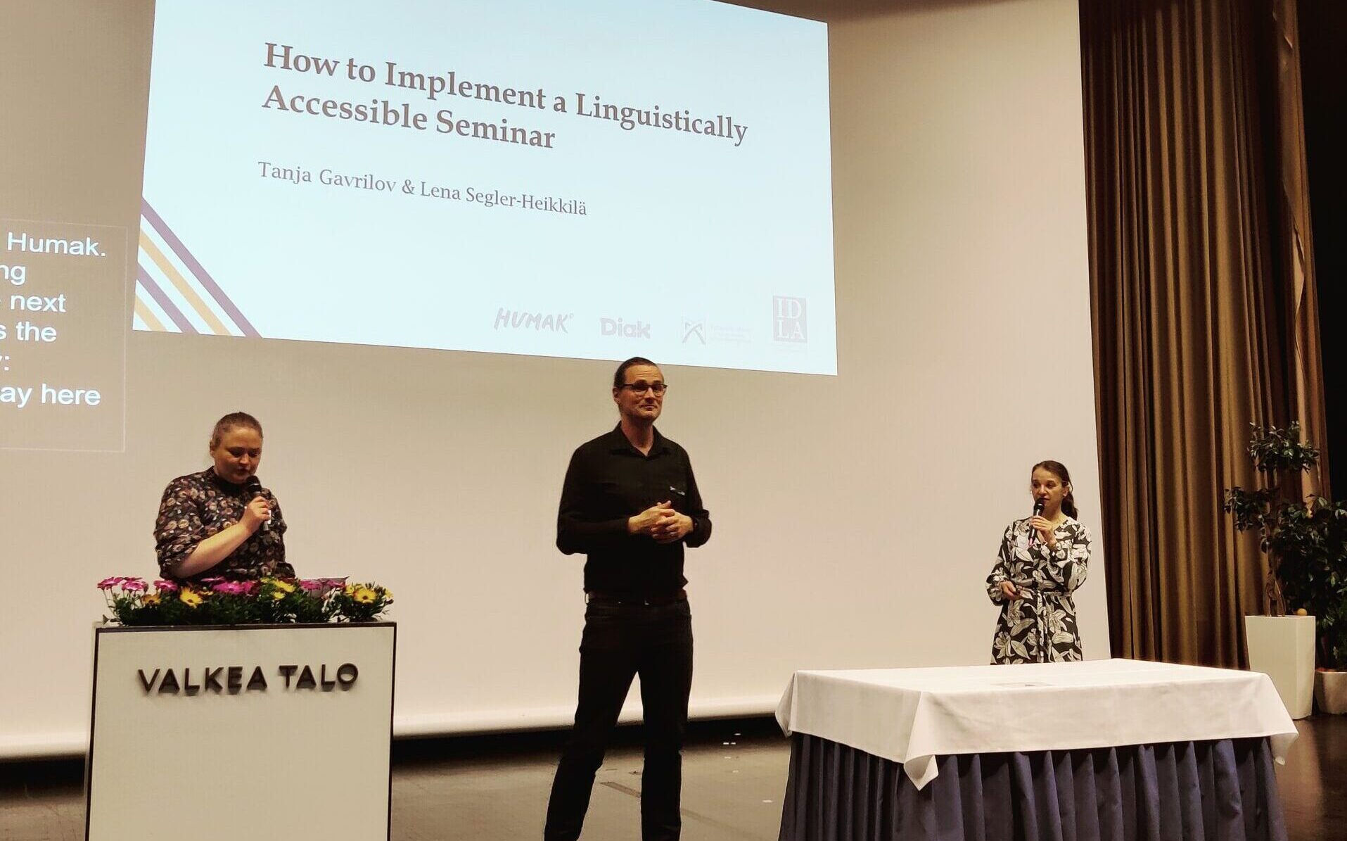 Tanja Gavrilov ja Leena Segler-Heikkilä seisovat Valkean talon auritoriossa. Heidän välissään on viittomakielen tulkki.