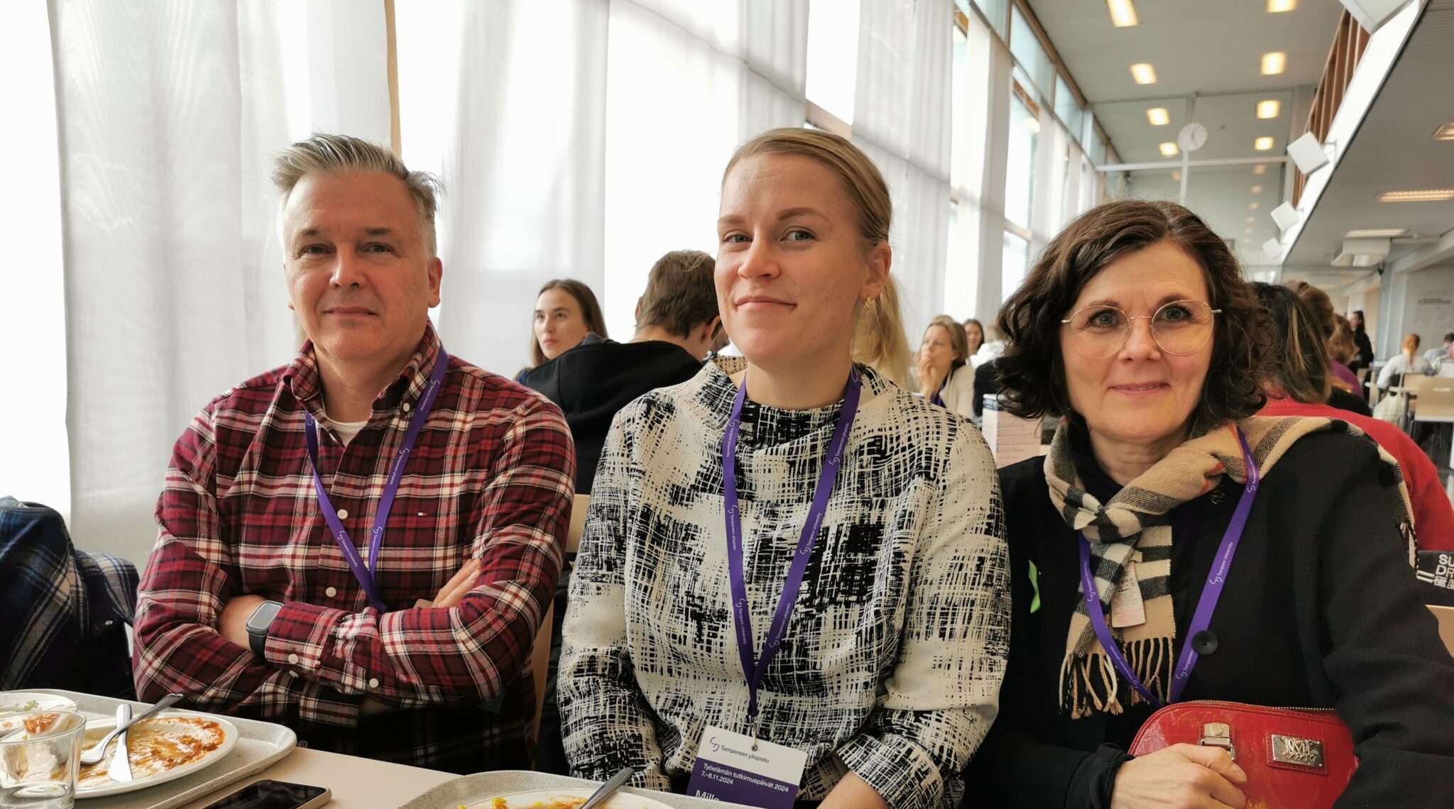 Kolme henkilöä istumassa ruokapöydän ääressä, kahdella edessä ruokalautanen. Taustalla opiskelijaruokala.