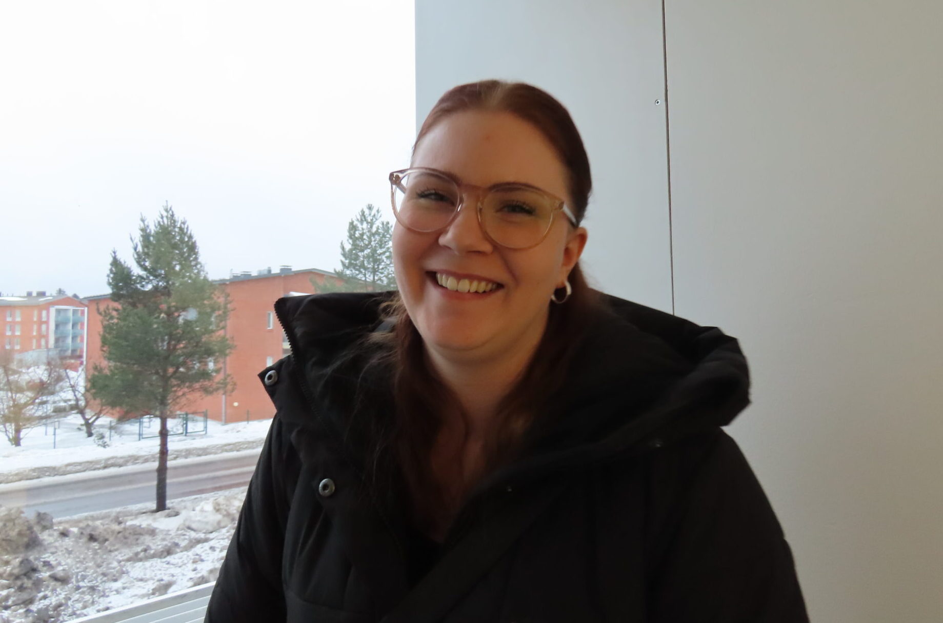 Nauravainen nainen kuvassa, taustalla lumista maisemaa, kerrostalon kulma, puu, ja sisätilakuvauksen seinä, kuvattavalla tukka kiinni, silmälasit, talvitakki.