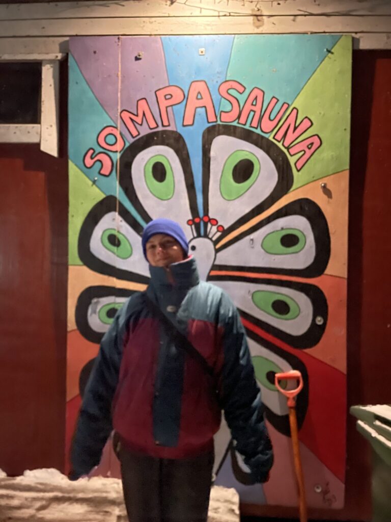 Woman smiling in front of Sompasauna public sauna's wall mural. The weather is cold and she is wearing a thick winter jacket, a beanie and gloves.