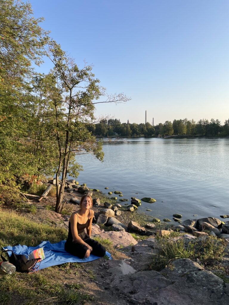 A young woman is sitting at the seafront in Helsinki. The weather is summery, the sky is blue and she is smiling at the camera.
