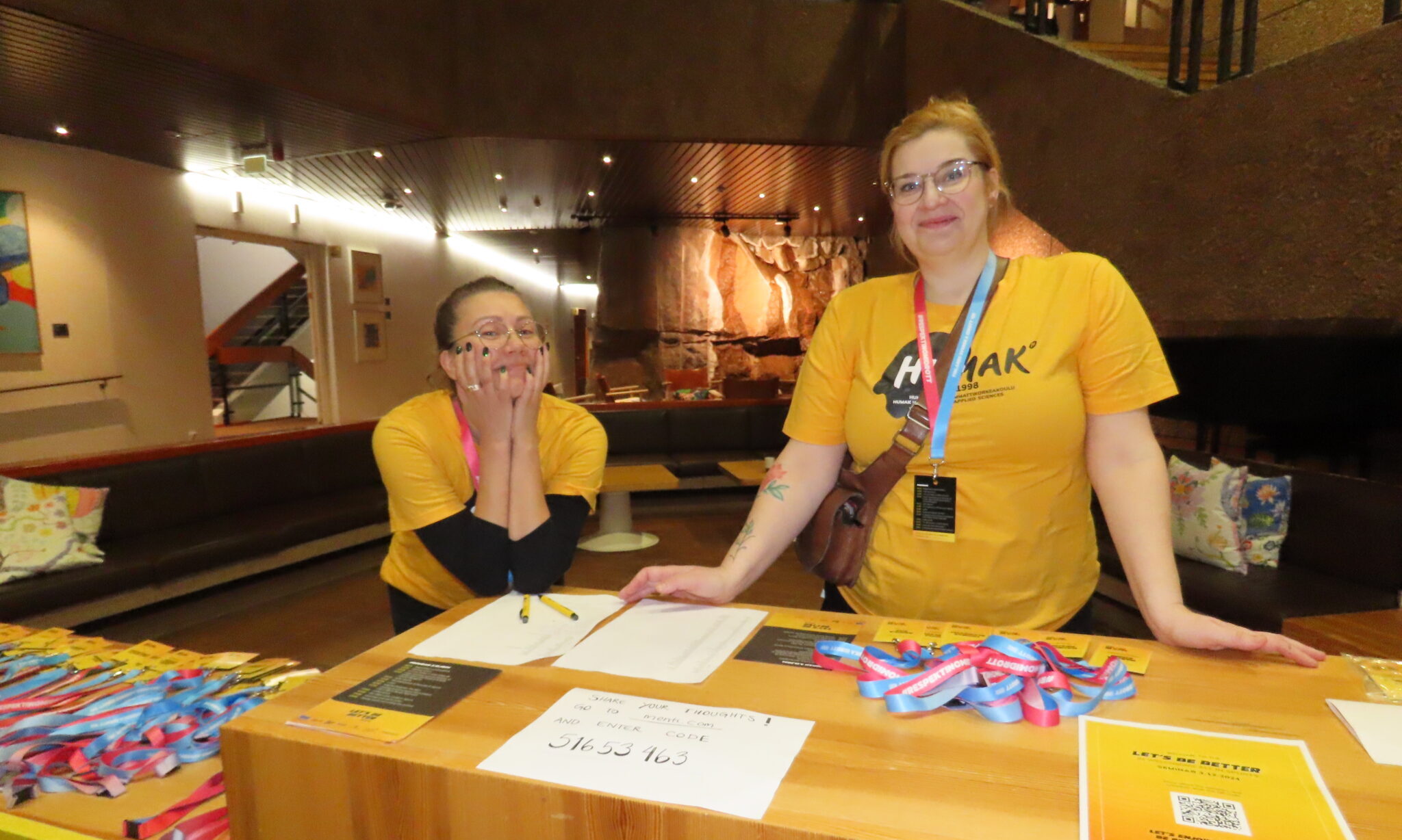 Kuvassa kaksi keltaisiin Humak paitoihin pukeutunutta naista, joista toinen nojaa kyynerpäillään pöytään ja toinen seisoo suorassa ja nojaa käsillään pöytään. Pöydällä on erilaista pikkutavaraa. 