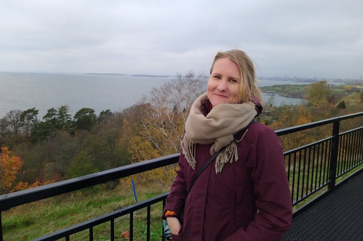 Luumun värinen takki, harmaa iso huivi, nainen ulkokuvassa, taustalla harmahtava merinäköala, syysmaisema. Ilse Lammila.