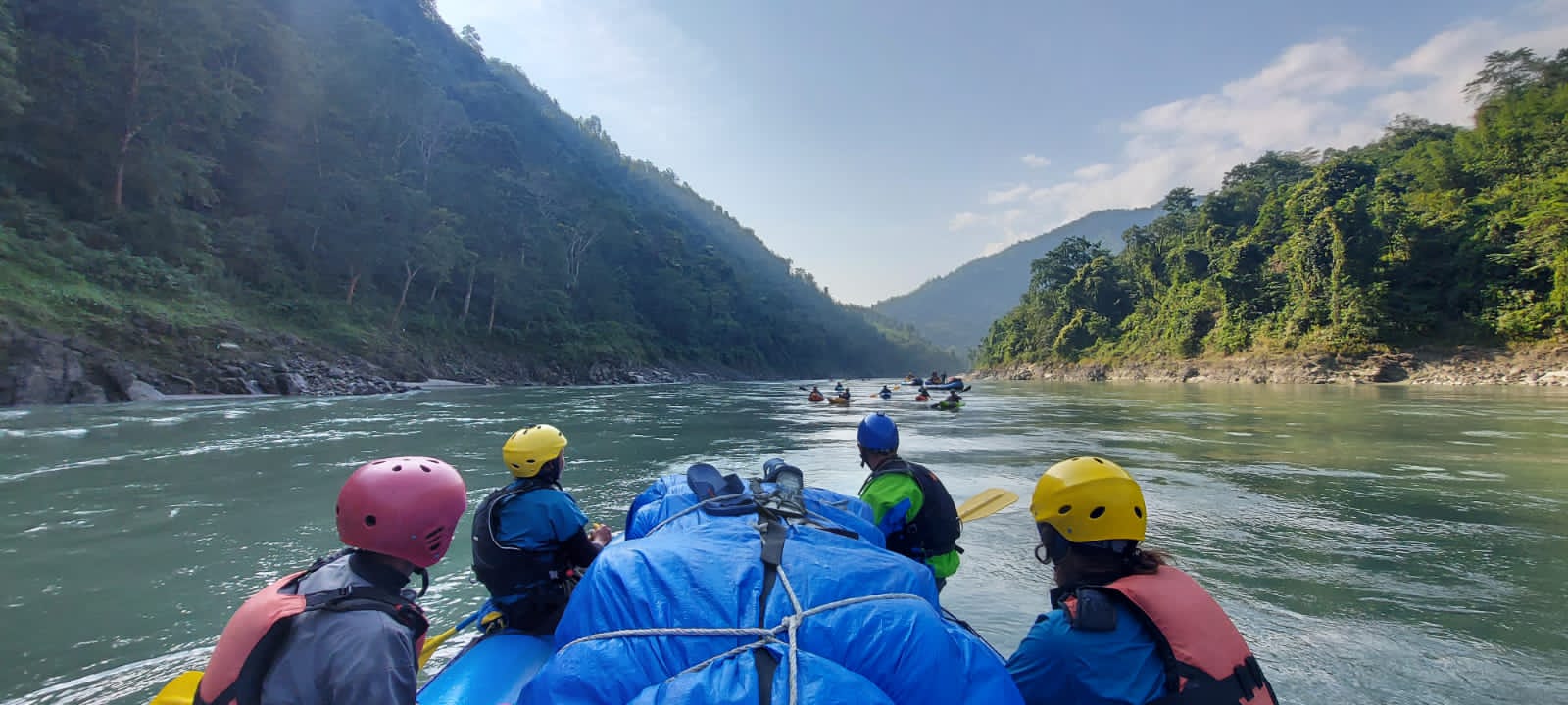 Several kayaks in different colours on a river with high shores with green trees.
