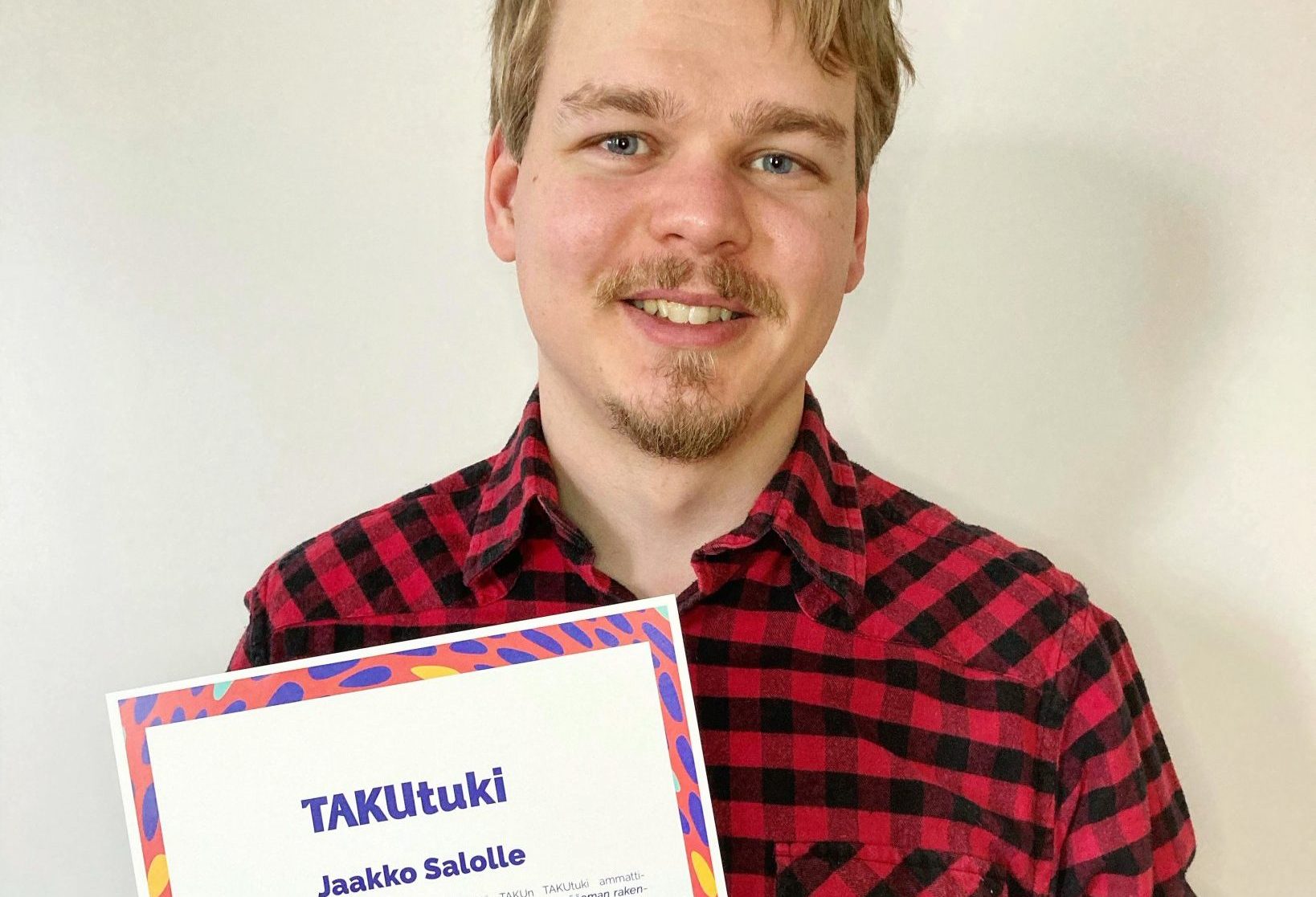 Hymyilevä mies mustapunaruutuisessa kauluspaidassa pitää kädessään stipendiä.