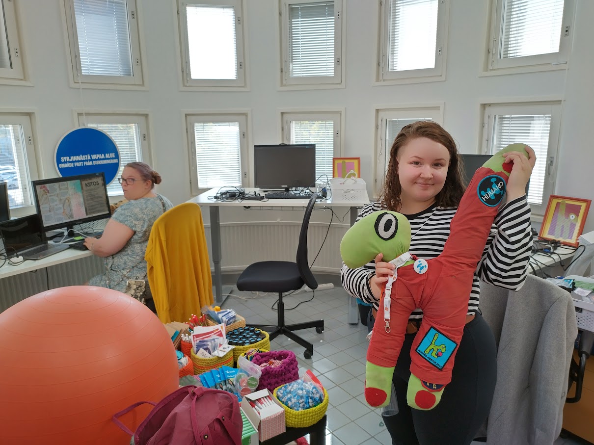 Kuvassa erilaisen pikkutavaran ja jumppapallon täyttämä toimistohuone, jossa taustalla pieniä neliömäisiä ikkunoita monessa rivissä. Kuvassa taustalla nainen istuu tietokoneen edessä ja on kääntynyt katsomaan takanaan olevaa kameraa, etualalla seisoo nainen. jonka sylissä on pehmomaskotti. 