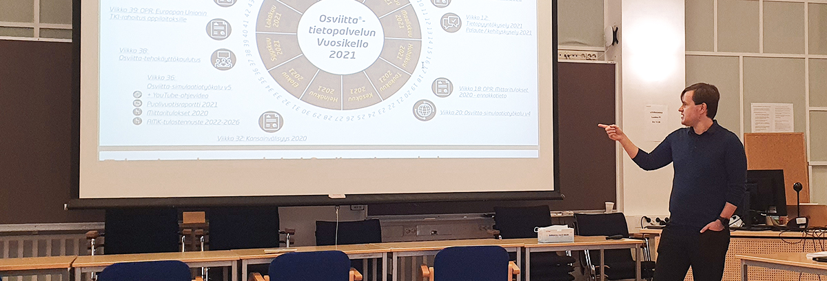 Ruskeahiuksinen mies (Jukka Jonninen) esittelee luokkahuoneessa Osviitta-tietopalvelun vuosikelloa, joka on heijastettu taustalla olevalle valkokankaalle.