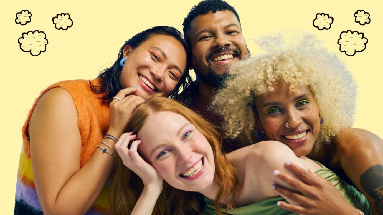 A group of four individuals standing close together against a yellow background with small cloud-like illustrations. They are posing in a friendly, casual manner, with some placing their hands on each other's shoulders.