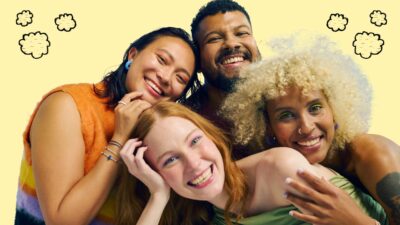A group of four individuals standing close together against a yellow background with small cloud-like illustrations. They are posing in a friendly, casual manner, with some placing their hands on each other's shoulders.