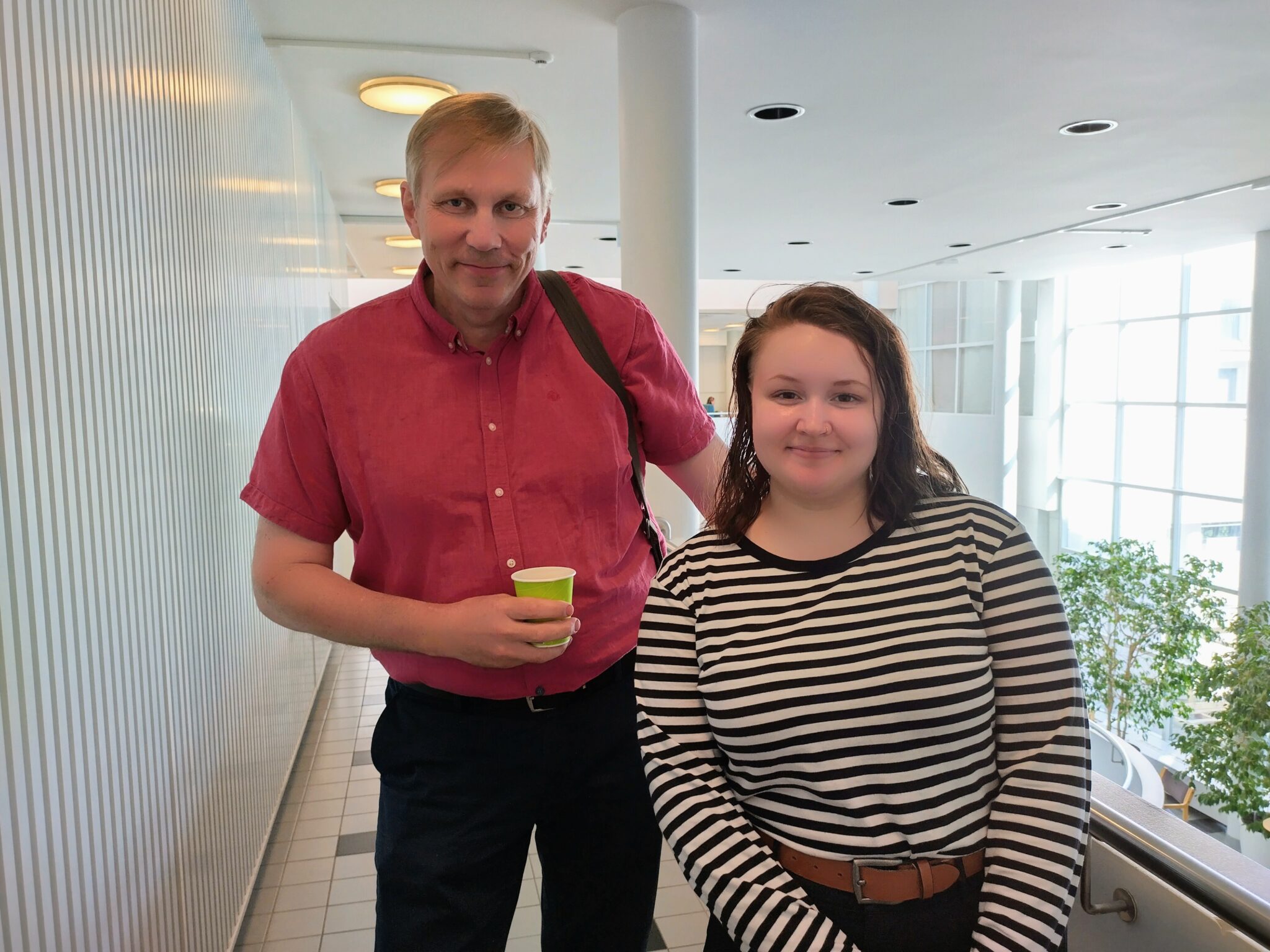 Kuvassa mies ja nainen käytävässä, yläpuolella valoja. Miehellä kädessään kahvimuki. 