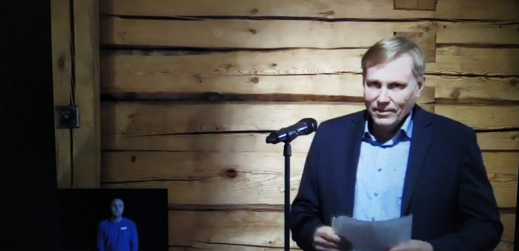 Jukka Määttä, mies tummassa takissa, sinisessä paidassa seisoo hirsiseinän edessä, mikrofonin takana, kädessä papereita, kuvakaappaus videokuvasta.