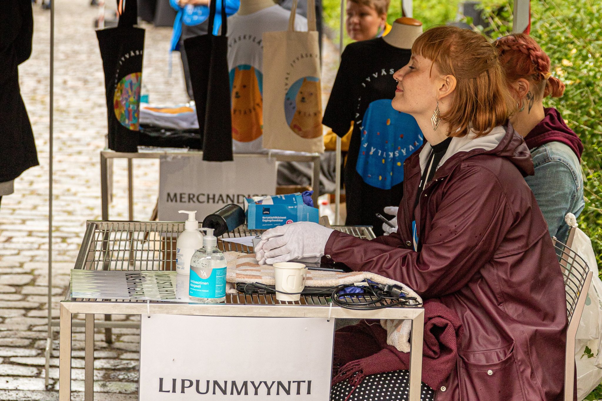 Kuvassa punahiuksinen nainen hymyilee ja istuu lipunmyyntipisteen vieressä.