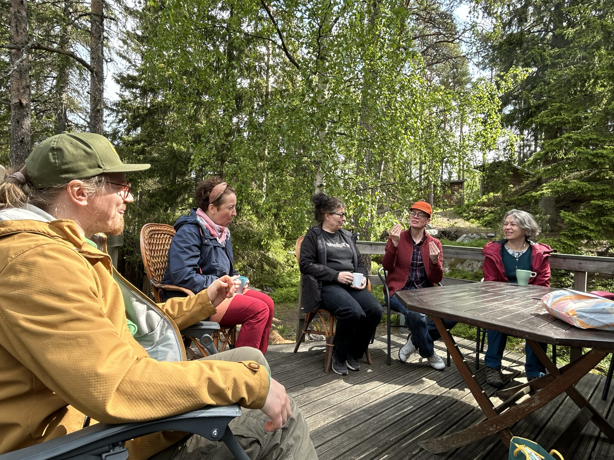 Viisi henkilöä istuu metsän keskellä terassilla ulkoiluvaatteet päällään. Yksi viittoo, ja muut katsovat häntä.