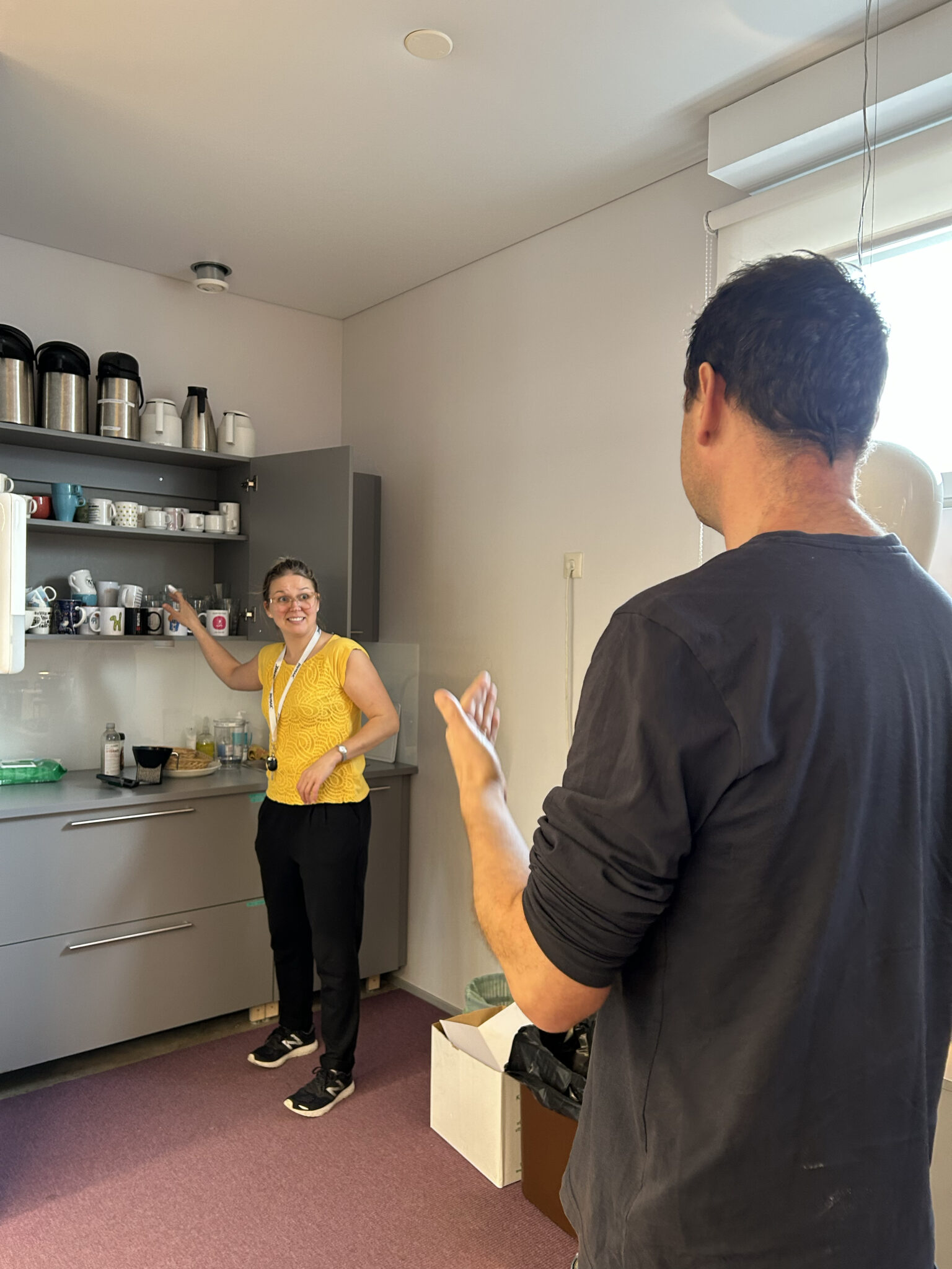 Vasemmalla oleva henkilö katsoo oikealla olevan henkilön viittomista ja yrittää samalla ottaa takanaan olevasta keittiön kaapista kuppia.