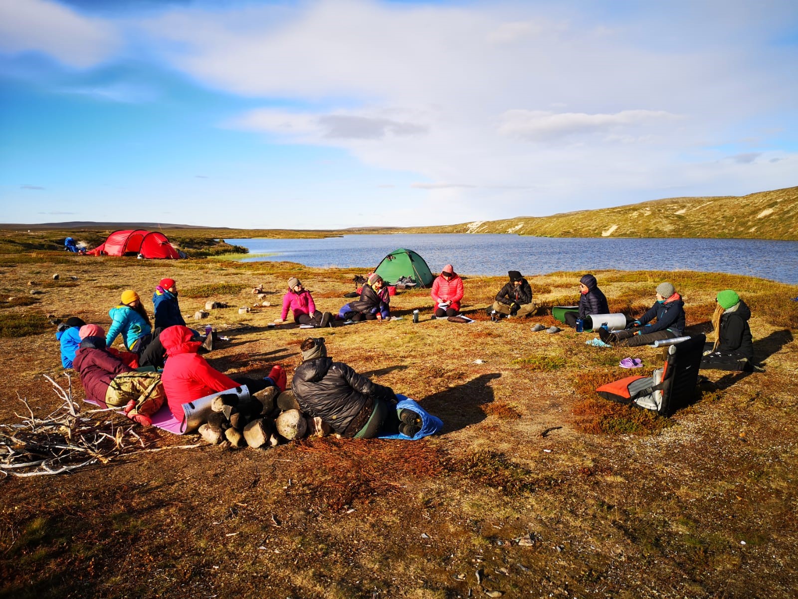 Ulkona ryhmä istuu levittäytyneenä pitämässä taukoa tai jotain, istuvat maassa, taustalla vesimaisemaa, sini-valkoinen taivat, ihmisillä ulkovaatteet ja taustalla punainen sekä vihreä teltta sekä kuvassa etualla risuja ja polttopuita.. 