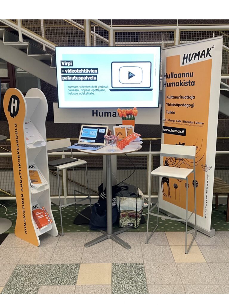 Kuvassa on Humakin esitetelineen ja roll-upin välissä esittelypöytä. Taustalla on näyttö, jossa on Virpi-videopalautustehtävän mainos.