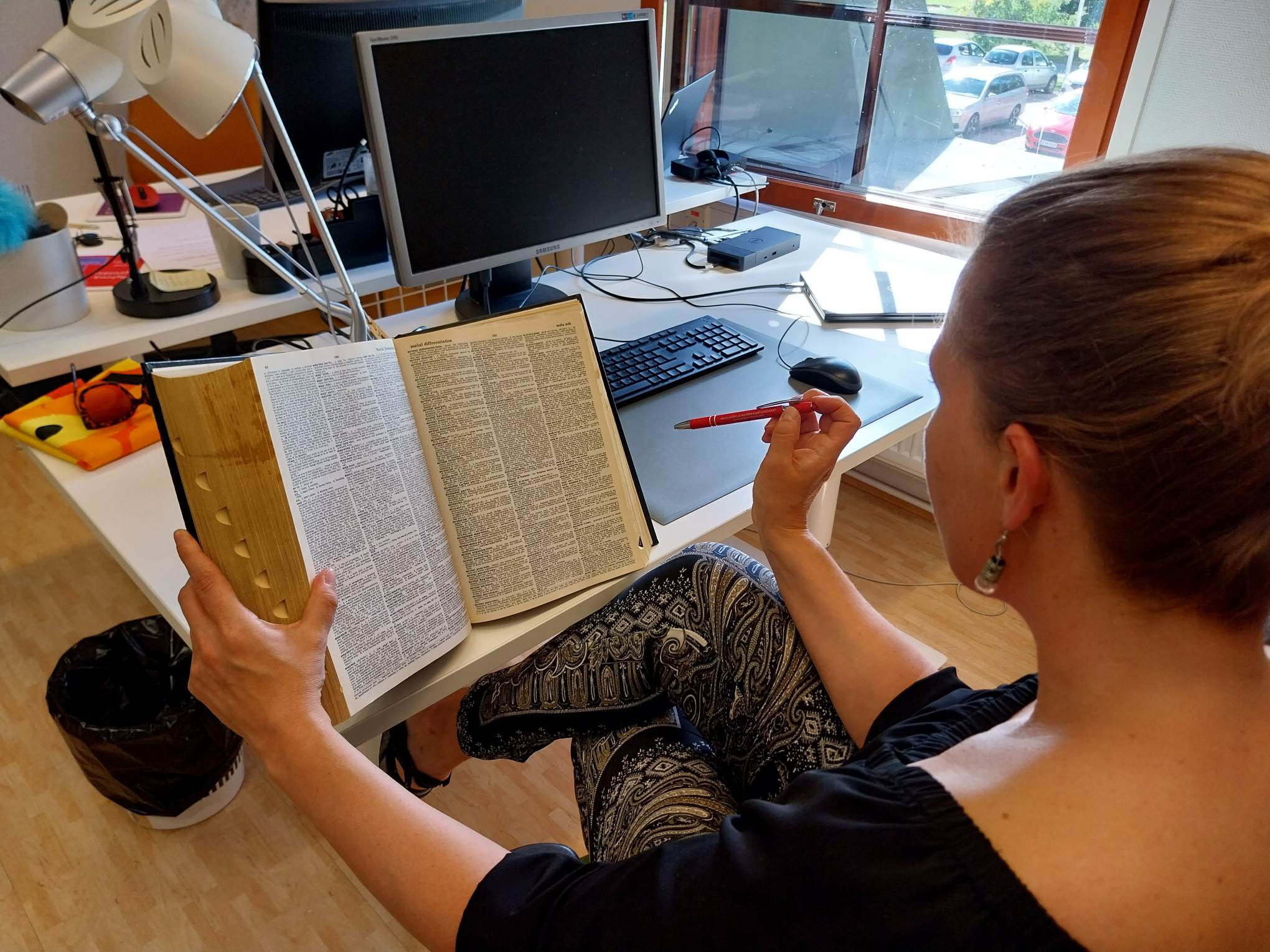 Nutturapäinen nainen istuu tietokonepöydän ääressä ja tutkii eräittäin suurikokoisen sanakirjan aukeamaa, joka on täynnä pientä tekstiä. Naisen oikeassa kädessä on kynä, joka osoittaa sanakirjan tekstin suuntaan.