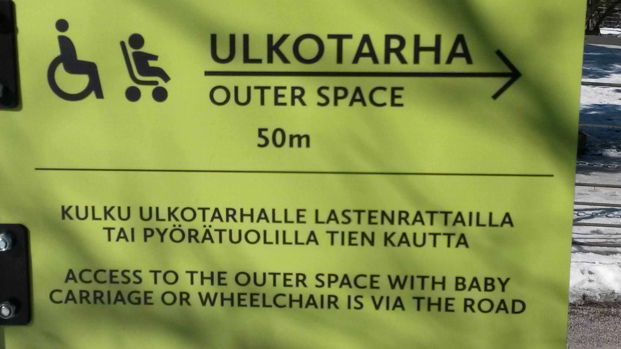 Keltainen kyltti, jossa mustat painatukset. Vasemmassa ylänurkassa pyörätuoli- ja lastenvaunusymbolit. Otsikon alla oikealle osoittava nuoli. Kaikki tekstit tikkukirjaimin. Otsikko: ”Ulkotarha – Outer Space”. Teksti: ”Kulku ulkotarhalle lastenrattailla tai pyörätuolilla tien kautta – Access to the outer space with baby carriage or wheelchair is via the road”.