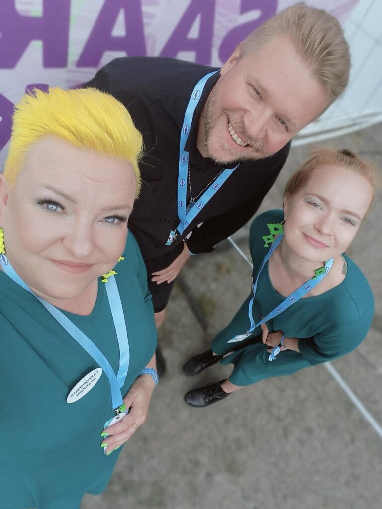 Kuvassa kolme henkilöä yläetukulmasta kuvattuna. Keskellä ylhäällä olevalla miehellä musta paita, kahdella naisella hänen molemmin puolin turkoosit mekot. Kaikki hymyilevät ja katsovat kameraan.