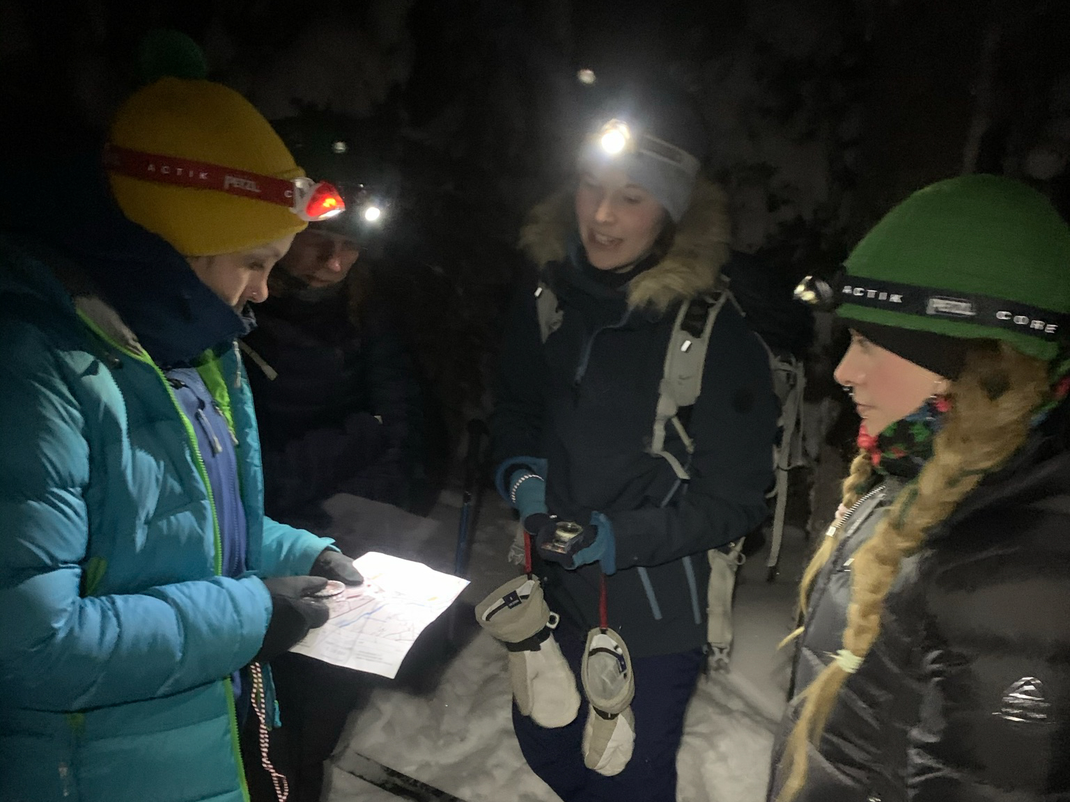 Kolme naista ulkona ulkovaatteissa seisoo ringissä. Yhdellä palaa otsalamppu ja toisella on kädessään kartta.
