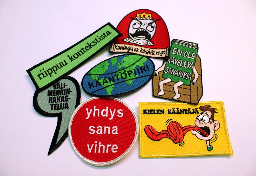 Seitsemän eri muotoista ja eri väristä kangasmerkkiä, joissa seuraavat tekstit: riippuu kontekstista; välimerkinrakastelija, kuningas on kauhea rage (kuva vihaisesta kuninkaasta valtaistuimella), kääntöpiiri (pohjakuvana maapallo), en ole kävelevä sanakirja, yhdys sana vihre [sic], kielen kääntäjä (kuvassa epätoivoinen mieshahmo, jonka pitkä kieli on solmussa). 