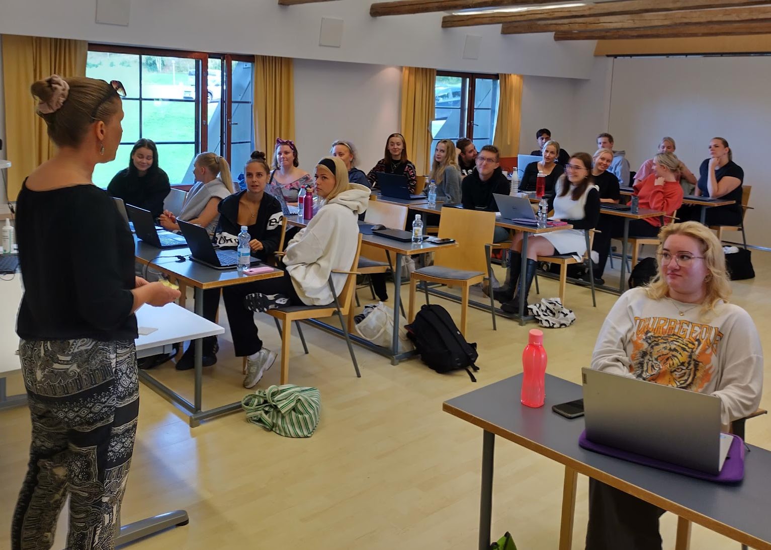 Luokan edessä selin kameraan naispuolinen, nutturapäinen opettaja mustassa paidassa ja kirjavissa housuissa. 19 opiskelijaa istuu luokassa neljän hengen pöytärivistöissä ja seuraa opetusta.