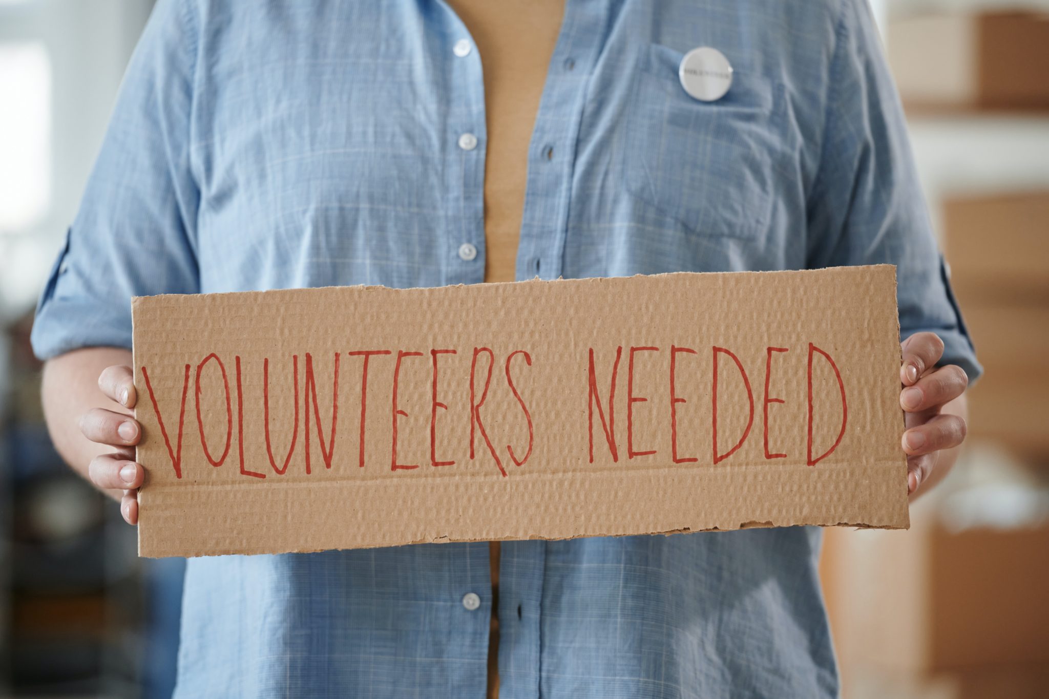 Henkilö pitelee pahvikylttiä edessään. Kyltissä lukee volunteers needed.
