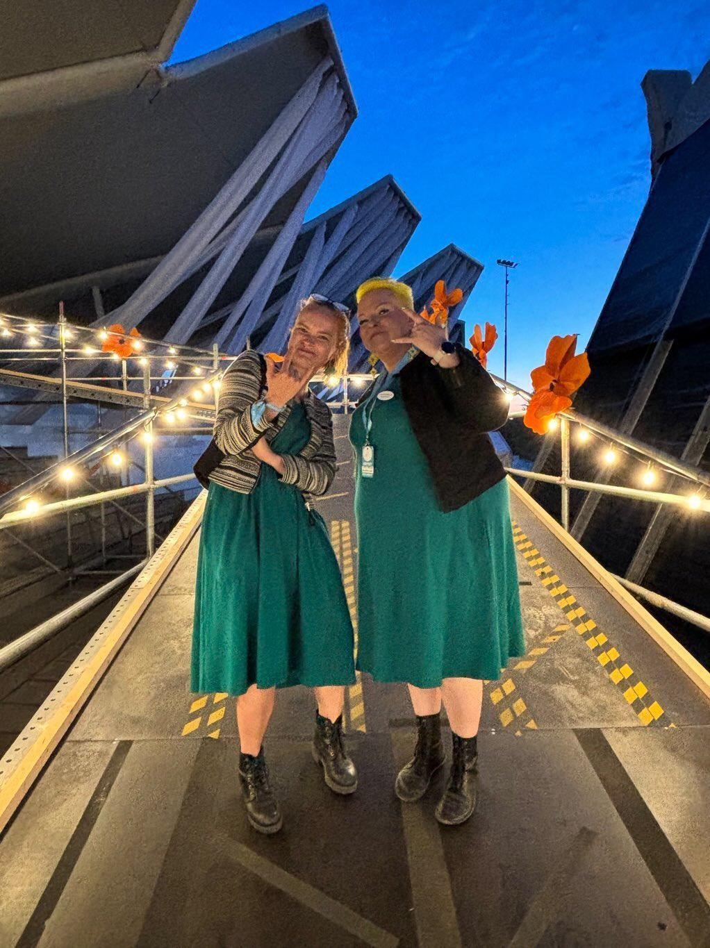 Kuvassa kaksi naista vihreissä mekoissa ja molemmat näyttävät kameralle rock-käsimerkkiä. Seisovat rampilla, jonka kaiteet on valaistu kausivaloilla. Taustalla näkyy tummansininen taivas. 