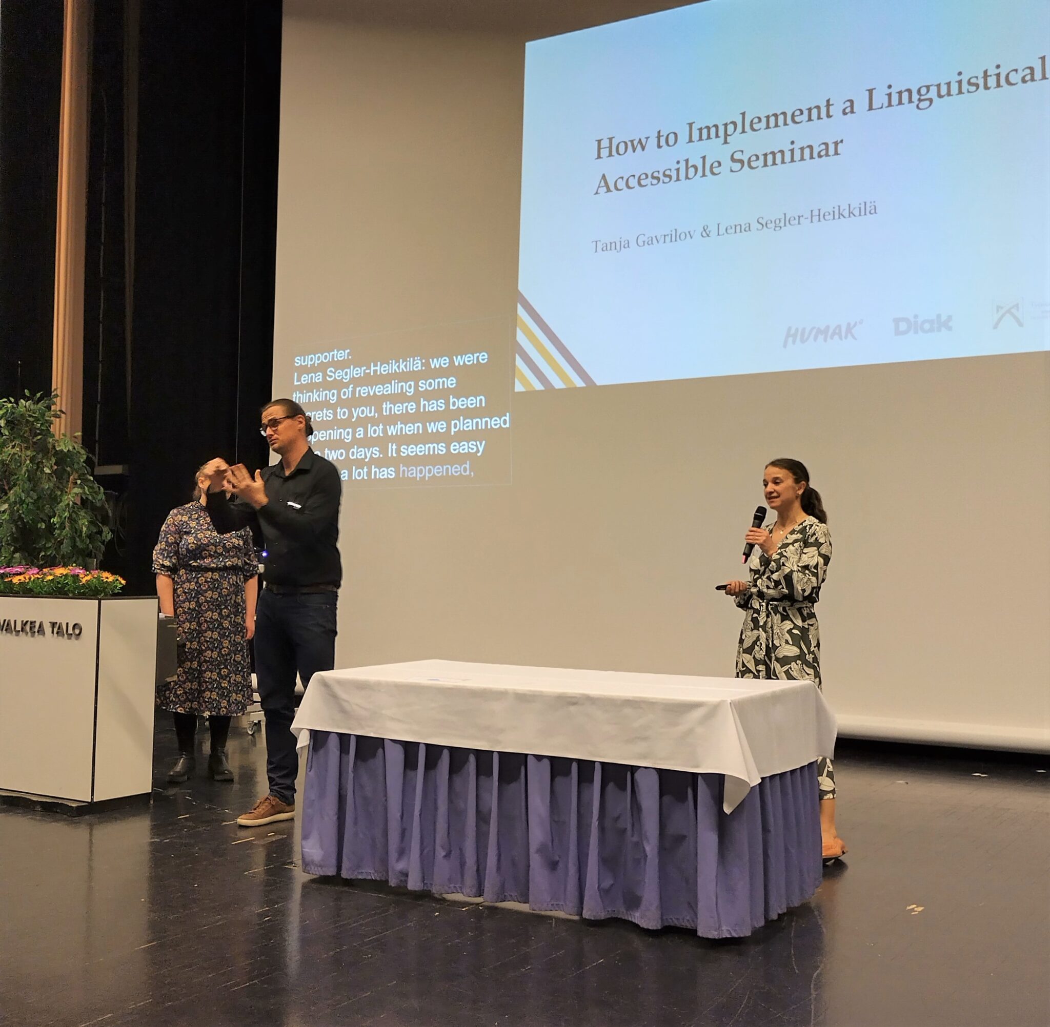 Kolme ihmistä seisoo suuren valkokankaan edessä, johon on heijastettu esitys. Yksi ihmisistä puhuu mikrofoniin ja yksi viittoo.