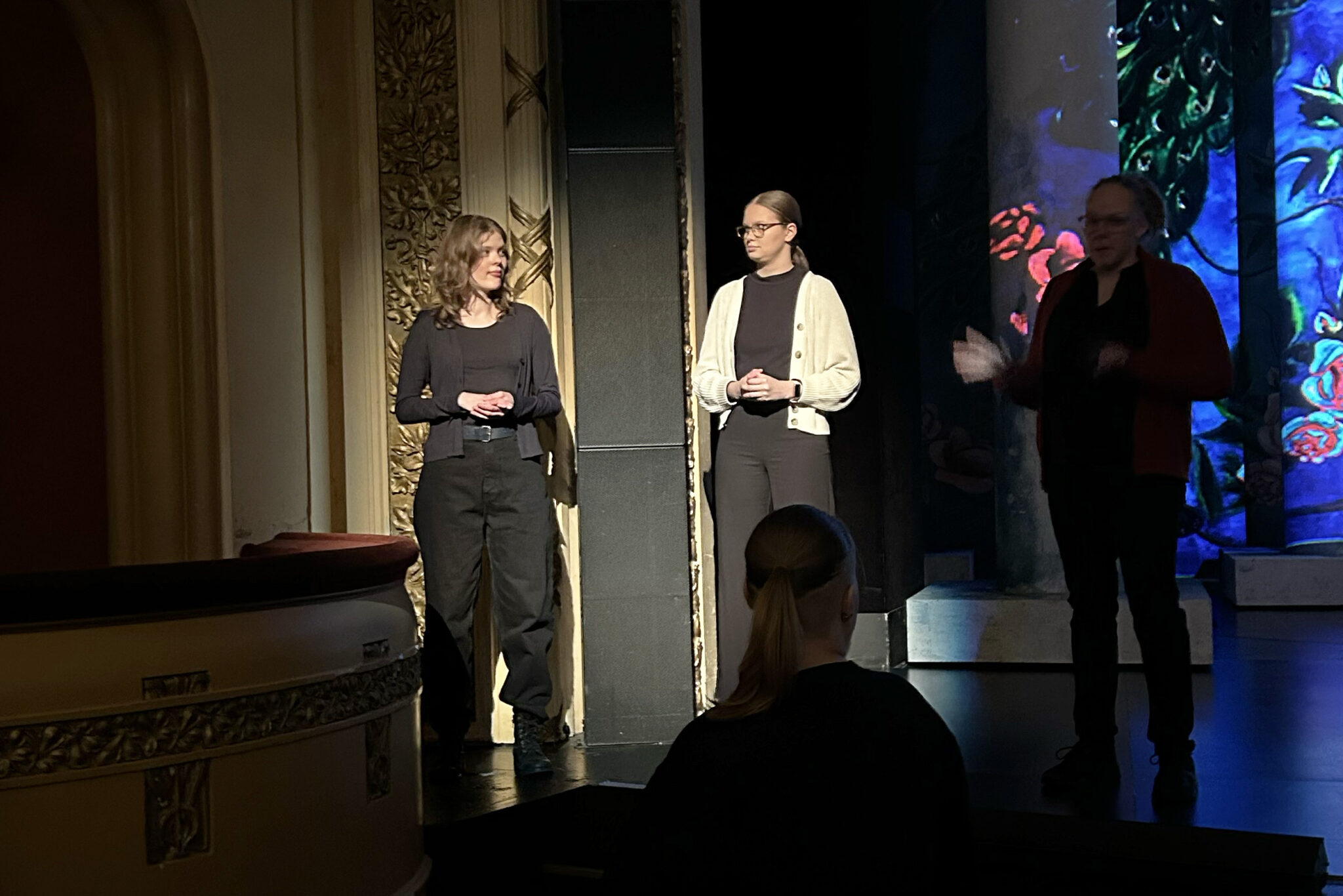 Kolme henkilöä seisoo teatterin lavalla. Kaksi heistä seisoo korkean mustan kaiuttimen molemmin puolin ja katsoo toisiaan. Heihin kohdistuu valo. Kolmas henkilö seisoo oikealla puolella pimeässä ja viittoo. Vasemmassa reunassa on aitio. Yksi henkilö seisoo lavan edessä selin kameraan ja katsoo lavalla olevia.