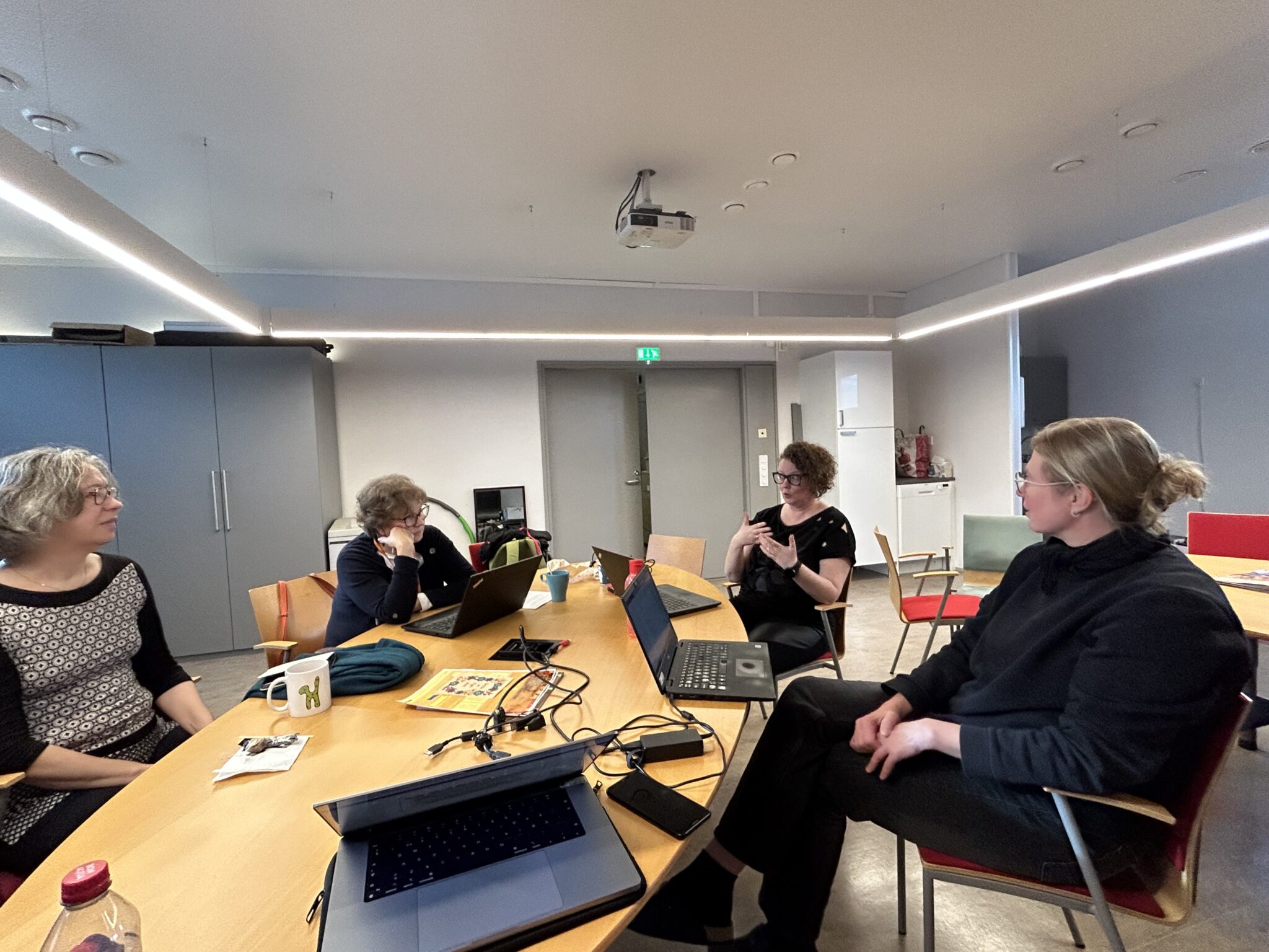 Neljä henkilöä istuu soikion muotoisen pöydän ympärillä keskustellen viittomakielellä