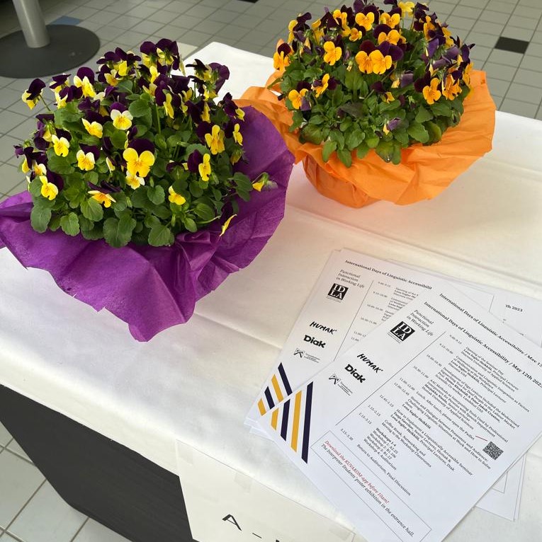 Neliön muotoinen värivalokuva. Kuvassa on pöytä, jolla on valkea liina. Pöydällä on kaksi orvokkiasetelmaa. Orvokkien värit ovat violetti ja oranssi. Kukkien lisäksi pöydällä on seminaarin käsiohjelmat. Käsiohjelmissa toistuu samat värit kuin kukissa. Ohjelmasta erottuu IDLA-seminaarin, Humakin, Diakin ja Työsuojelurahaston logot.