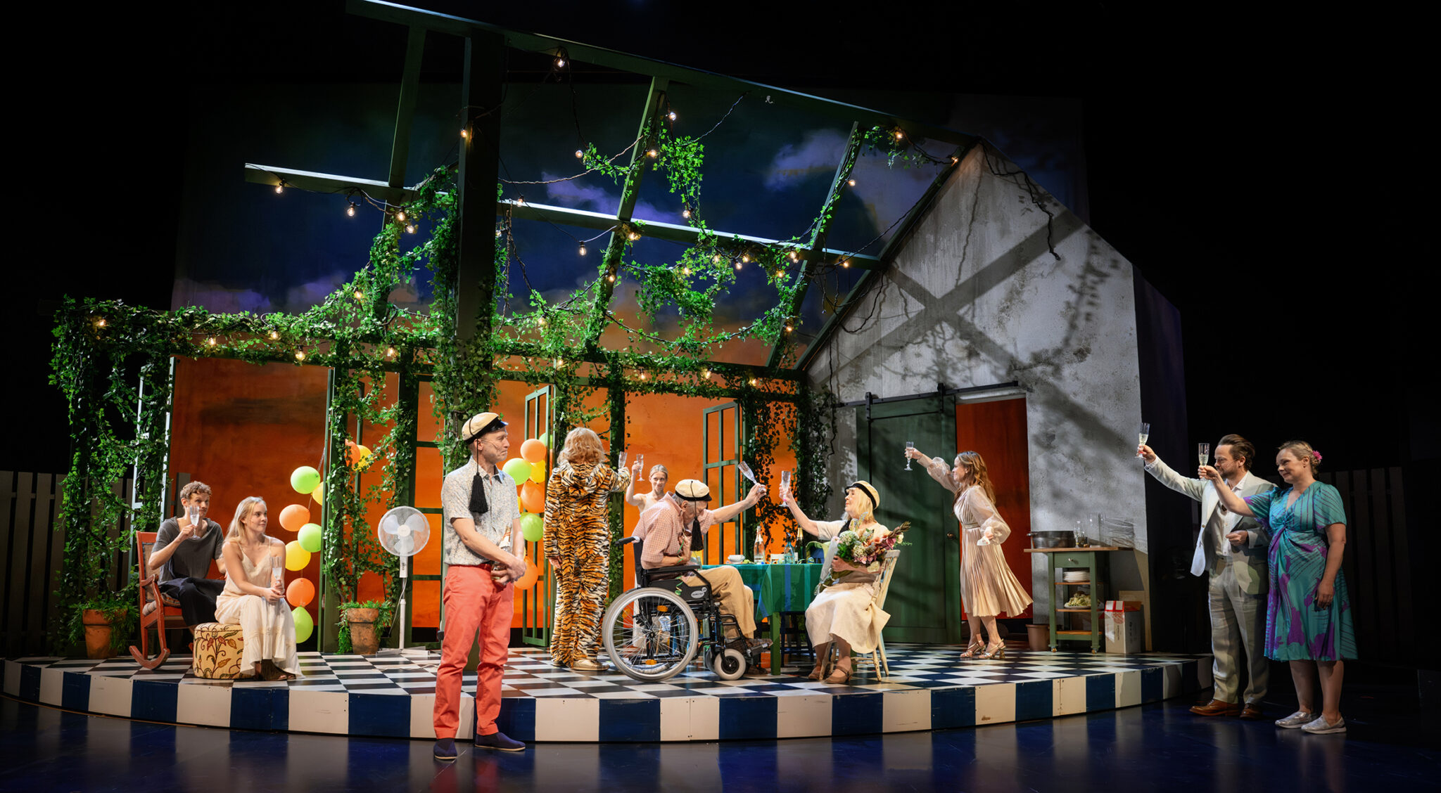 Kuvassa on kymmenen henkilöä teatterin lavalla. Lavasteena on talon seinä ja pergola, jossa on köynnöksiä ja juhlavalosarja sekä ilmapalloja. Henkilöt ovat juhlavissa vaatteissa, paitsi yhdellä on yllään tiikeriasu. Osalla on päässään ylioppilaslakki. Osa seisoo, osa istuu. Keskellä kuvaa pöydän ääressä on vanha mies pyörätuolissa ja vanha nainen kukkakimppu sylissään. Suurin osa kohottaa kuohuviinilasia.