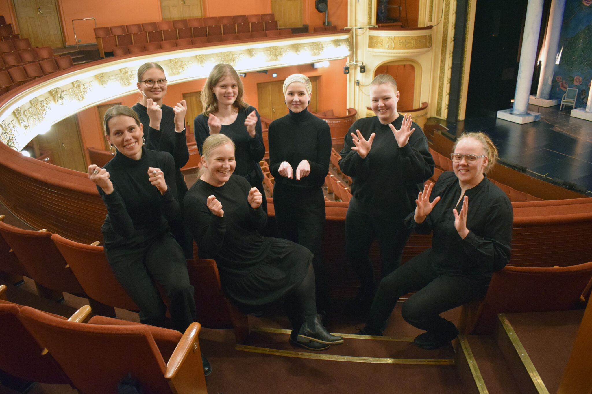 Seitsemän henkilöä on teatterin ensimmäisellä parvella kaiteen vieressä selkä lavalle ja permannolle päin. Takarivissä neljä henkilöä seisoo, eturivissä kolme istuu. Vasemmalla puolella olevat viittovat viittomaa JUHLA, oikealla puolelle kaksi viittoo viittomaa ELÄMÄ ja keskellä oleva viittoo viittomaa SIINÄ-SIVUSSA. Kaikki katsovat kameraan ja hymyilevät.