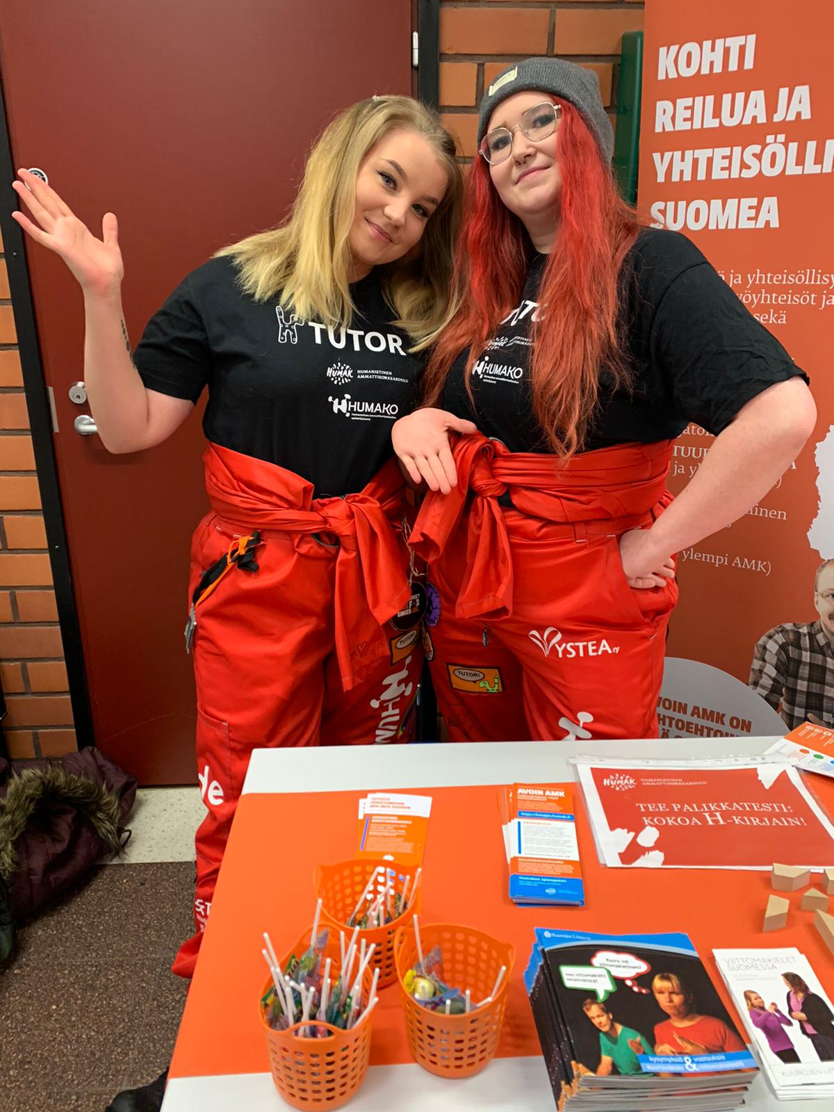 Kaksi hymyilevää opiskelijatyttyöä seisovat messupöydän takana päällään punaiset opiskelijahaalarit.