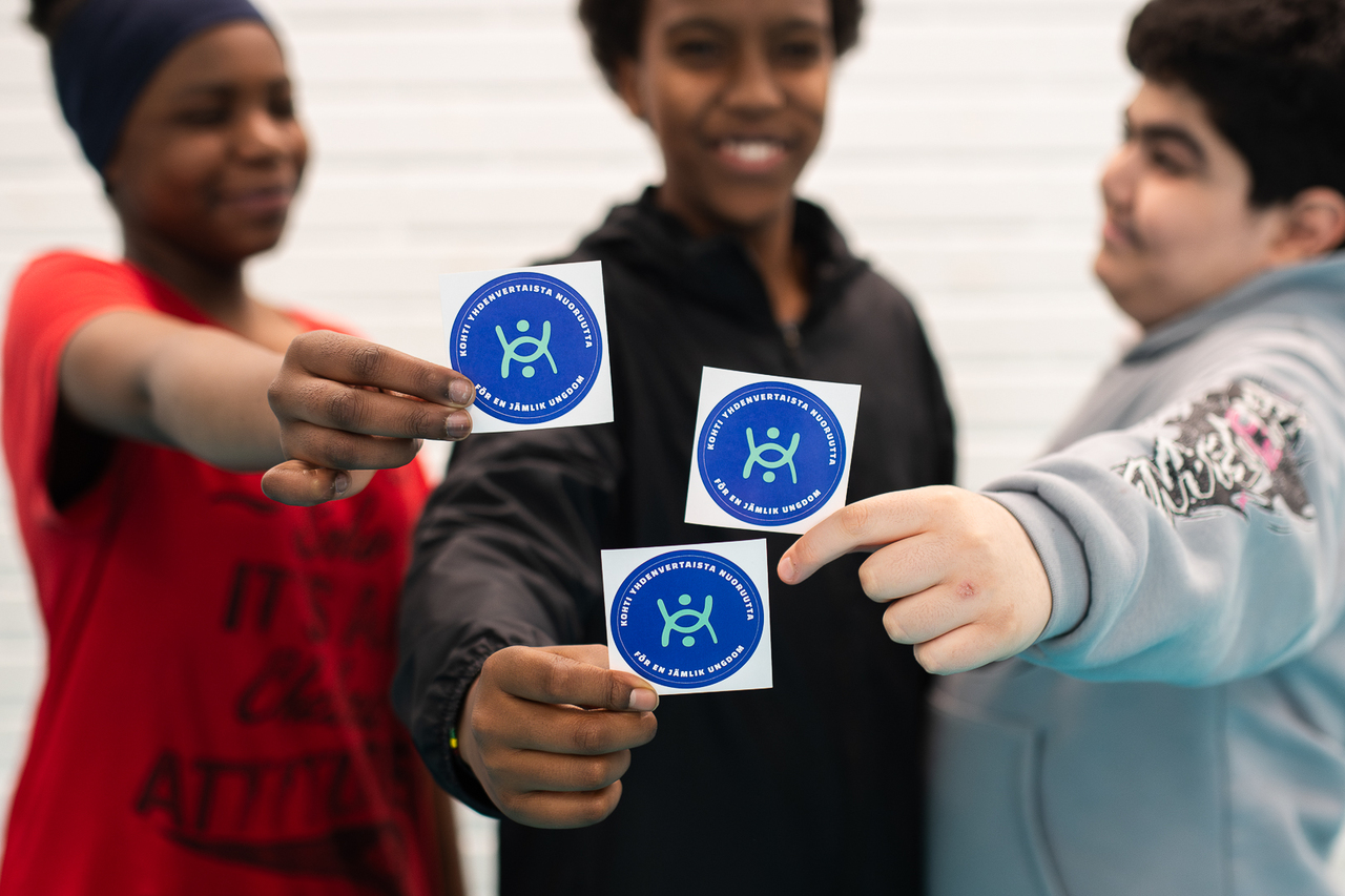 Nuoret pojat näyttävät kameralle hymyillen tarroja, joissa on Yhdenvertaisuuden nuorisoalan osaamiskeskuksen logo.