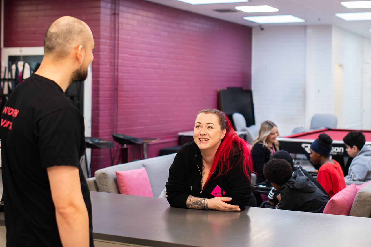 Nainen, jolla on kirkkaanpunaiset hiukset, nojaa hymyillen tiskiin ja juttelee miehen kanssa. Taustalla sohvilla ja pöydän ääressä istuu nuoria sekä toinen aikuinen, ja huoneessa on biljardipöytä. Seinät ovat vaaleanpunaisia ja valkoisia.