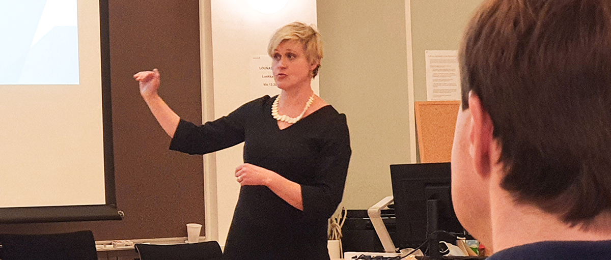 Vaaleahiuksinen nainen (Ida Mielityinen) puhuu luokkahuoneen edessä. Kuvassa etualalla tummahiuksinen mies, selkä kuvaajaa kohti.