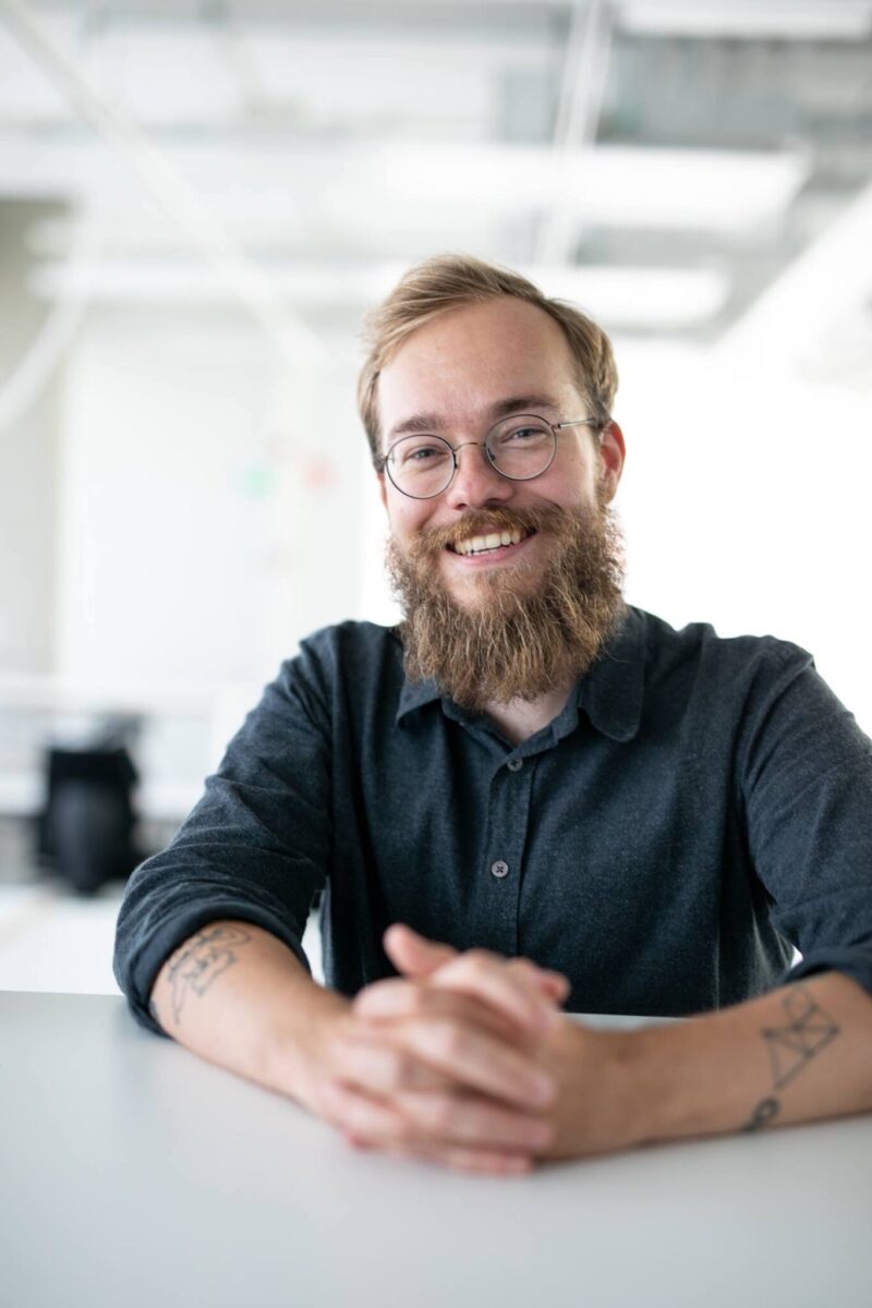 Ruskeapartainen mies pitää käsiään pöydällä ja nojaa siihen. Hän hymyilee ja hänellä on silmälasit.