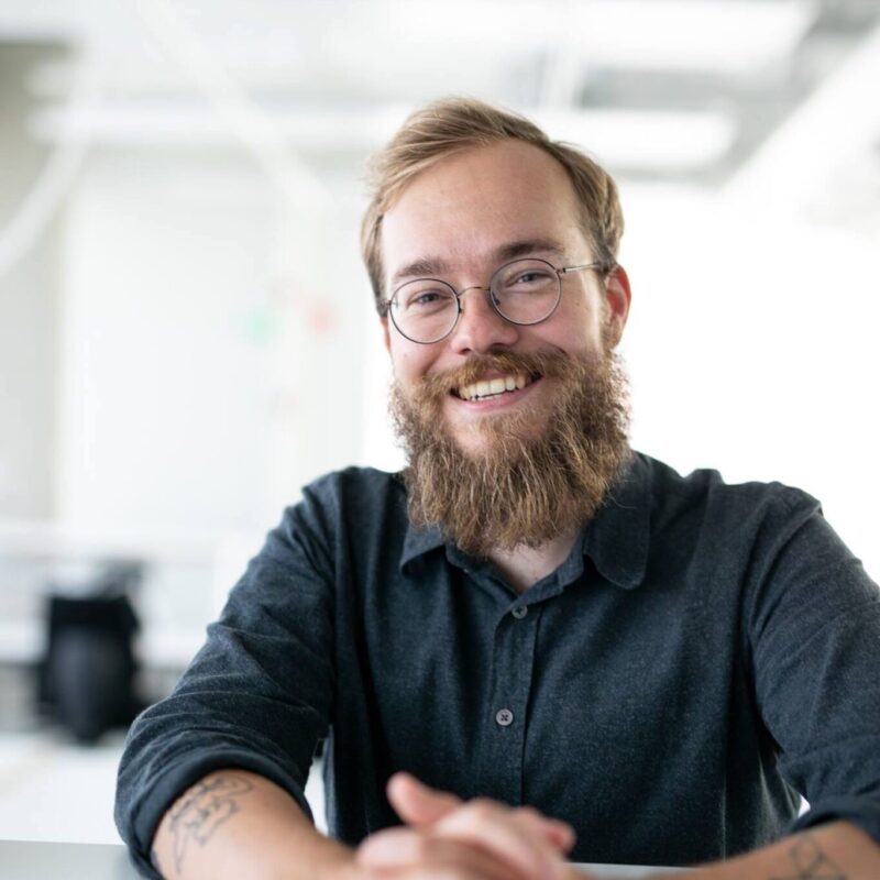 Ruskeapartainen mies pitää käsiään pöydällä ja nojaa siihen. Hän hymyilee ja hänellä on silmälasit.