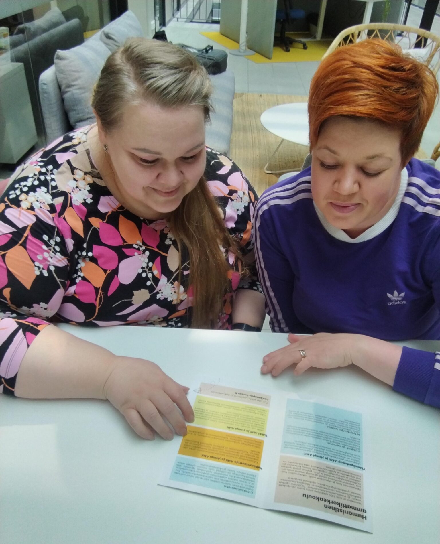 Kuvassa kaksi naista istuu vierekkäin pöydän ääressä ja tutkivat paperista esitettä. 