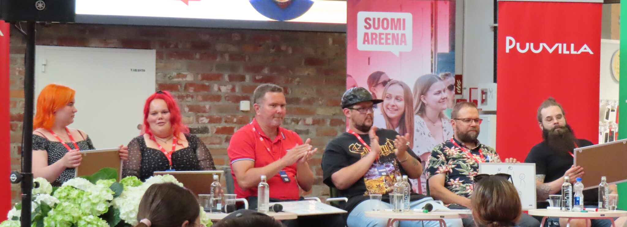 Kuvassa SuomiAreenan paneeli, jossa viisi osallistujaa istuu vierekkäin lavalla. Vasemmalta lähtien kaksi naista, kolme miestä sekä puhujanpöntön takana naisjuontaja. Edustalla taka-alalta kuulijoiden päitä ja kukkia. 
